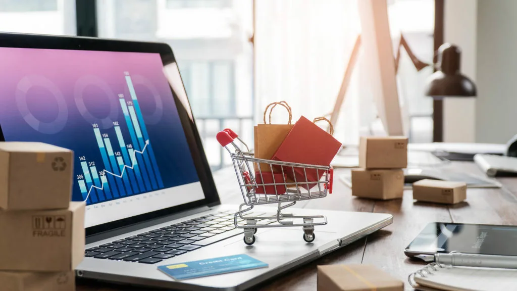 An open laptop with a tiny shopping cart perched about the mousepad.