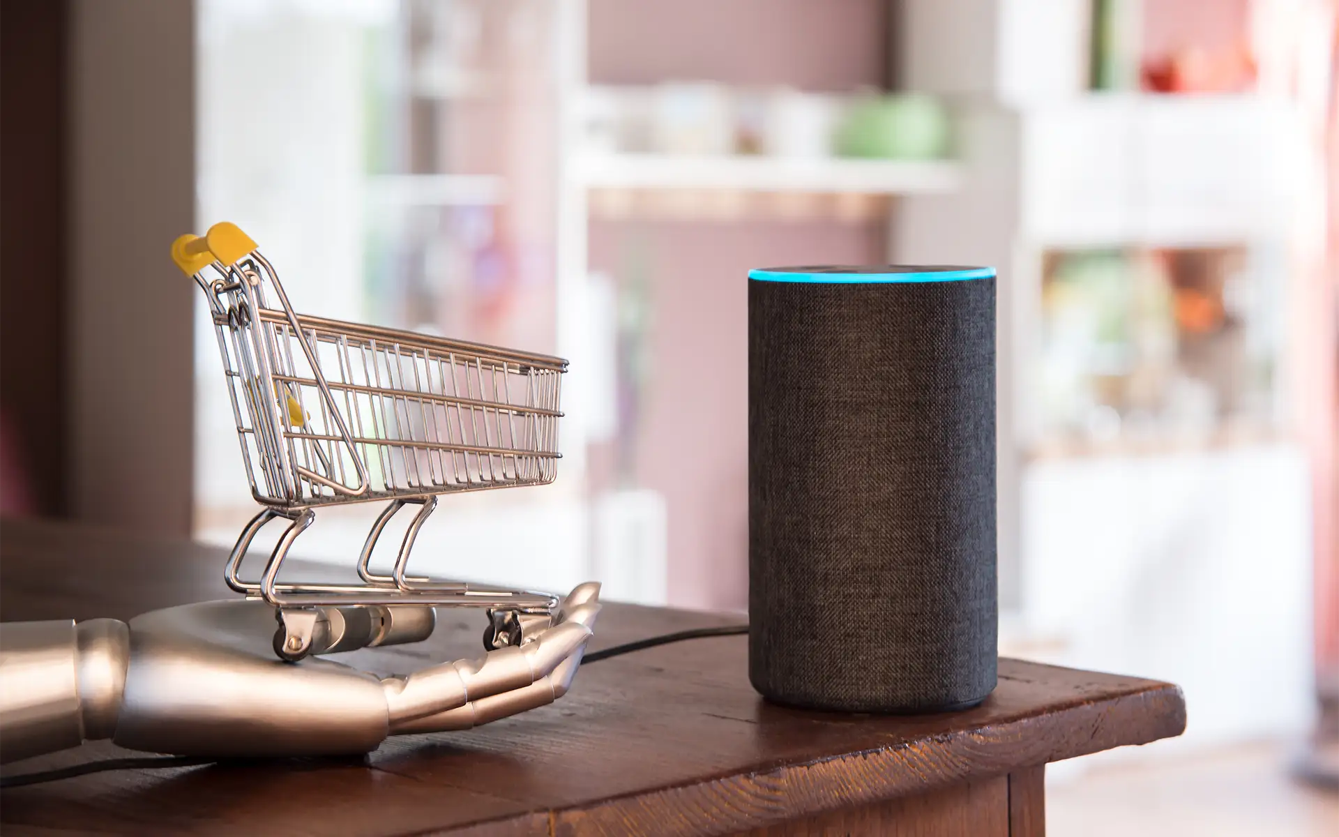 A miniature shopping cart sitting in a robotic hand next to a smart speaker with voice recognition to signify eCommerce AI voice shopping