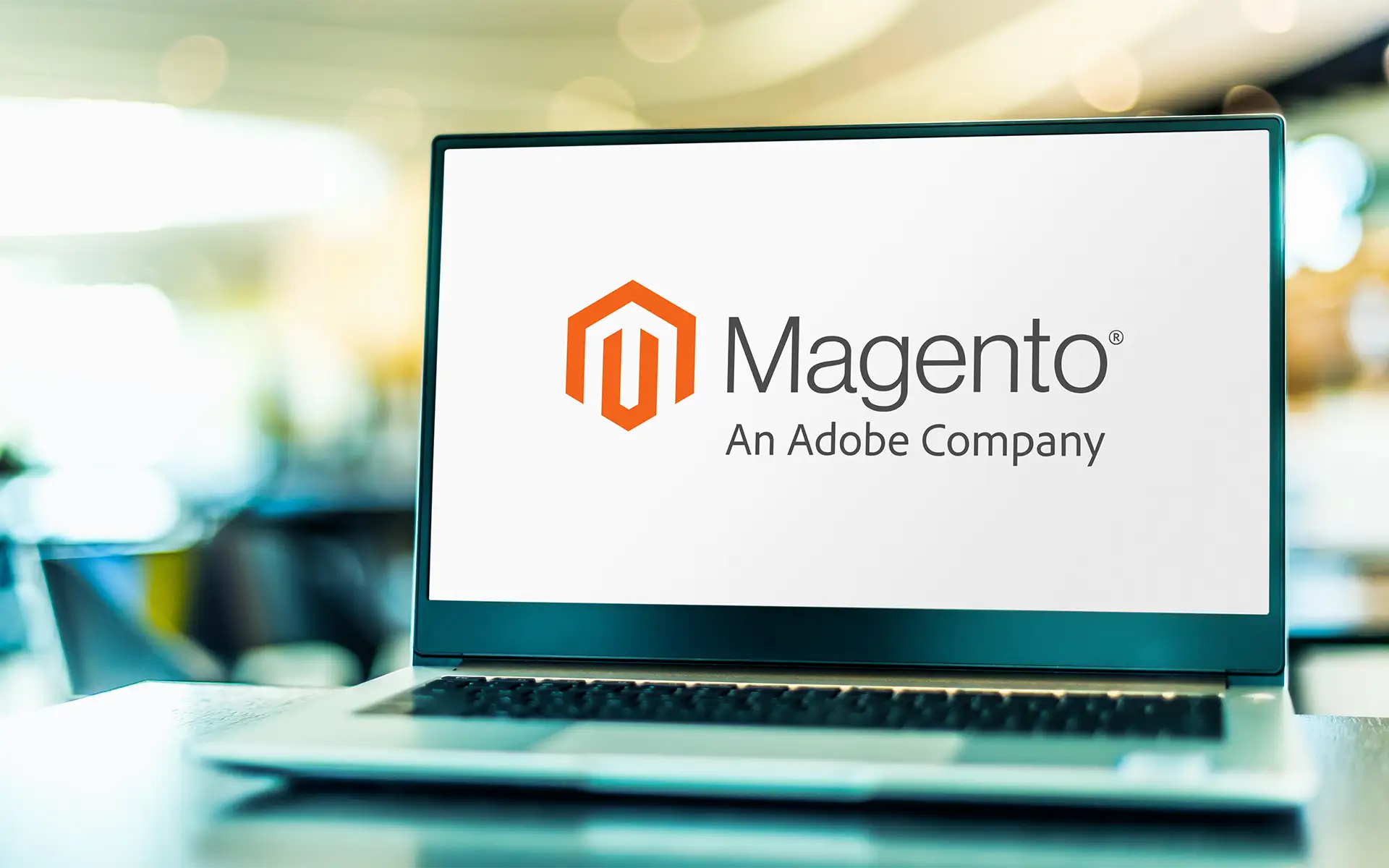 An open laptop sittin gon a table with the words "Magento-An Adobe Company" on it's screen, next to the Magento logo